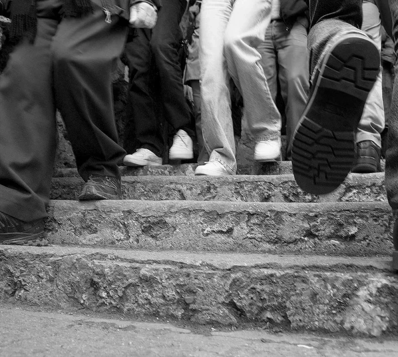 Pascale · 21/03/2026 · 1000 x 897 Cette photographie en noir et blanc capture principalement des **pieds et jambes** de plusieurs personnes descendant ou montant des **escaliers usés**. Le **sujet principal** est l’interaction entre les individus et l’architecture dégradée.La **composition** est dynamique, avec un **cadrage serré** sur les pieds et les marches, créant un effet de mouvement. La **lumière** semble naturelle mais contrastée, accentuant les textures et ombres. Les **couleurs dominantes** sont les gris et noirs, renforçant l’aspect urbain et vieilli.Le **style** est documentaire et réaliste, évoquant une **ambiance** de quotidien, de fatigue ou de précarité, avec une touche de mélancolie.