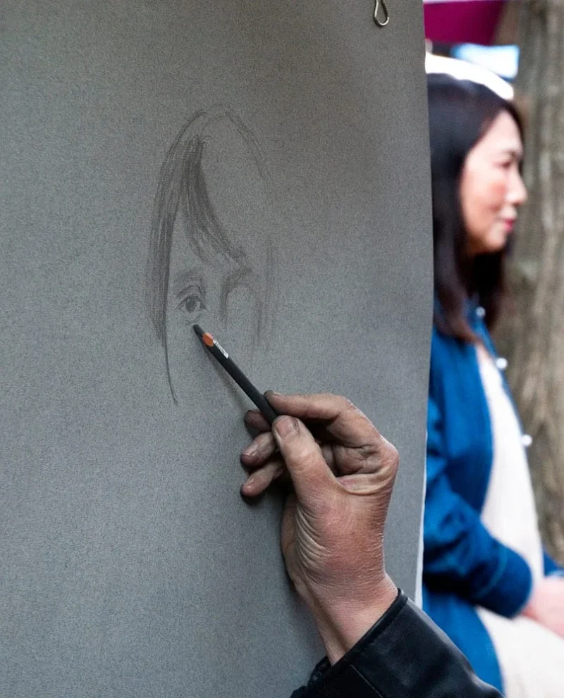 Pascale · 21/03/2026 · 807 x 1000 Cette photographie montre une personne en train de dessiner un portrait au crayon sur une feuille posée contre un mur. Le sujet principal est le dessin en cours et la main tenant le crayon.La composition est simple et centrée, avec un cadrage serré sur le geste artistique. La lumière est naturelle et douce, mettant en valeur les traits délicats du dessin. Les couleurs dominantes sont les tons gris (crayon) et bleutés (vêtements de la personne en arrière-plan).L’ambiance est calme et concentrée, évoquant la patience et la précision du travail artistique.