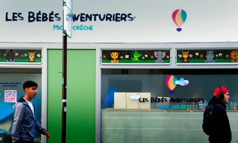 Pascale · 22/03/2026 · 1000 x 603 Cette photographie montre l’entrée d’une crèche nommée **« Les Bébés Venturiens »**. Le sujet principal est un homme masqué marchant devant l’établissement.La composition met en valeur l’enseigne lumineuse et colorée, avec des ballons et des motifs joyeux. Le cadrage est serré sur l’entrée, avec une perspective légèrement basse. La lumière est naturelle et claire, accentuant les couleurs vives : bleu, vert, rouge et jaune dominent.Le style est urbain et moderne, évoquant une ambiance chaleureuse et accueillante pour les jeunes enfants.