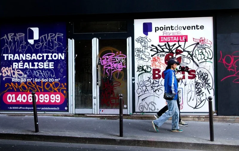 Pascale · 15/03/2026 · 1000 x 632 Cette photographie montre un homme marchant devant un magasin dont la vitrine est taguée et affichée avec des inscriptions.Le sujet principal est l’homme en mouvement. La composition inclut des éléments urbains : graffitis, affiches ("Transaction réalisée" avec un numéro de téléphone) et enseigne "point de vente".Le cadrage est serré sur la vitrine et l’homme, avec une lumière naturelle diffuse. Les couleurs dominantes sont les tons froids (bleu, gris) et les touches de violet et noir des graffitis. L’ambiance est urbaine, réaliste et légèrement délabrée, reflétant une atmosphère de ville animée et artistique.