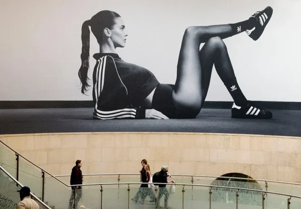 Pascale CHESNOT · 07/03/2026 · 1000 x 694 Cette photographie montre une **murale géante** représentant une athlète en position de pompe sur les mains, vêtue d’un short et d’un haut Adidas. La **composition** met en valeur le contraste entre le sujet dynamique et l’architecture urbaine en arrière-plan.Le **cadrage** est serré sur le mural, avec des personnes en bas qui donnent l’échelle. La **lumière** est naturelle et uniforme, sans ombres marquées. Les **couleurs dominantes** sont le noir, blanc et gris, renforçant un style épuré et moderne. L’**ambiance** est à la fois sportive et urbaine, évoquant énergie et mouvement dans un cadre contemporain.