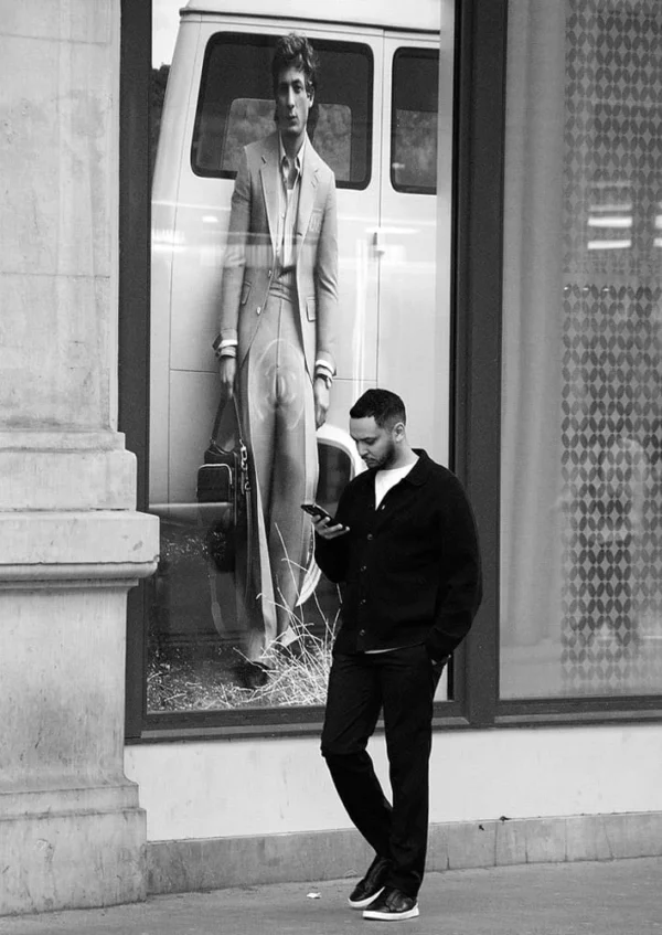 Pascale CHESNOT · 10/03/2026 · 708 x 1000 Cette photographie en noir et blanc met en scène un homme comme sujet principal, marchant sur un trottoir tout en regardant son téléphone. La composition oppose son mouvement à la statue géante reflétée dans une vitrine, créant une symétrie visuelle.Le cadrage est serré sur l’homme, avec la statue en arrière-plan. La lumière est naturelle, contrastée, accentuant les ombres et les détails. Les tons dominants sont les gris et noirs, renforçant une ambiance urbaine et intemporelle. Le style est documentaire, évoquant une scène du quotidien avec une touche artistique. L’ambiance est à la fois moderne et mélancolique.