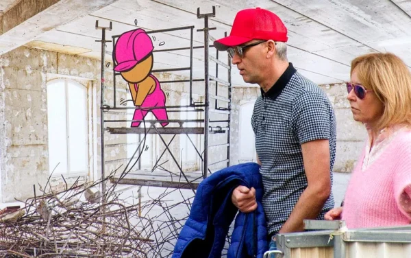 Pascale CHESNOT · 07/03/2026 · 1000 x 626 Cette photographie montre un couple assis sur un rebord de fenêtre ou une corniche. Le sujet principal est l’homme, vêtu d’un polo à carreaux et d’un chapeau rouge, observant une fresque murale représentant un personnage stylisé. La femme, à ses côtés, porte un haut rose et des lunettes.La composition est simple et naturelle, avec un cadrage serré sur les deux personnages. La lumière semble naturelle, douce et légèrement diffuse. Les couleurs dominantes sont les tons pastel (rose, bleu) et les contrastes vifs (rouge du chapeau, bleu du polo).Le style est réaliste et spontané, créant une ambiance détendue et presque intimiste.