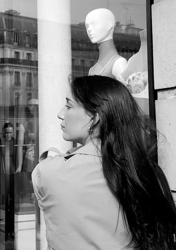 Pascale CHESNOT · 07/03/2026 · 637 x 904 Cette photographie en noir et blanc met en scène une femme comme sujet principal, vue de profil, les cheveux longs et le regard tourné vers la gauche. La composition joue sur la transparence d’une vitrine, reflétant une mannequin en arrière-plan. Le cadrage est serré sur la femme, avec une profondeur de champ qui isole son visage et ses épaules.La lumière est douce, naturelle, créant des contrastes subtils. Les tons dominants sont les gris et noirs, renforçant une ambiance introspective et élégante. Le style est sobre et minimaliste, évoquant une atmosphère à la fois urbaine et contemplative.