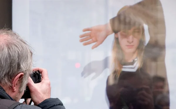 Pascale CHESNOT · 07/03/2026 · 1000 x 618 Cette photographie montre deux sujets principaux : un homme âgé prenant une photo avec un appareil argentique et une jeune femme reflétée dans un miroir ou une surface vitrée.La composition joue sur la superposition des images, créant un effet de profondeur. Le cadrage est serré sur les visages et les mains, mettant l'accent sur l'action de photographier. La lumière semble naturelle et douce, éclairant les sujets de manière uniforme. Les couleurs dominantes sont des tons neutres (beige, gris) avec des touches de brun et de noir, renforçant une ambiance sobre et intemporelle. Le style est documentaire et spontané, évoquant une scène du quotidien.