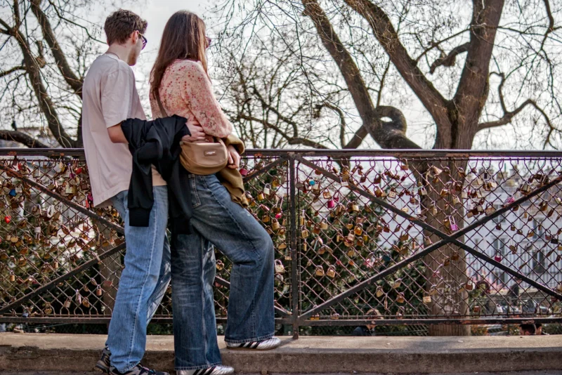 Frédéric · 21/03/2026 · 2517 x 1680 Cette photographie montre deux jeunes marchant main dans la main sur un pont. Le sujet principal est ce couple, dont les visages sont floutés, créant une ambiance intimiste.La composition est simple et naturelle, avec un cadrage serré sur les deux personnages. L’arrière-plan, composé de barrières métalliques et d’arbres sans feuilles, suggère un environnement urbain ou semi-urbain.La lumière est douce et naturelle, probablement en fin de journée. Les couleurs dominantes sont les tons neutres (gris, beige) et quelques touches de couleurs vives apportées par leurs vêtements (orange, bleu).Le style est réaliste et spontané, évoquant une ambiance calme et romantique.