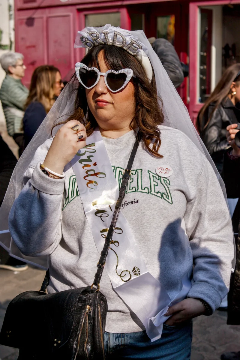 Frédéric · 21/03/2026 · 1120 x 1680 Cette photographie met en scène une femme comme sujet principal, vêtue d’un sweat à capuche et coiffée d’un voile de mariée stylisé, avec des lunettes à paillettes. Le cadrage est serré, centré sur elle, avec un arrière-plan flou montrant une rue animée.La lumière est naturelle et douce, mettant en valeur les détails de son visage et de ses accessoires. Les couleurs dominantes sont les tons pastel (blanc, gris) et des touches de rose et bleu. Le style est décalé et festif, évoquant une ambiance ludique et créative, presque carnavalesque.
