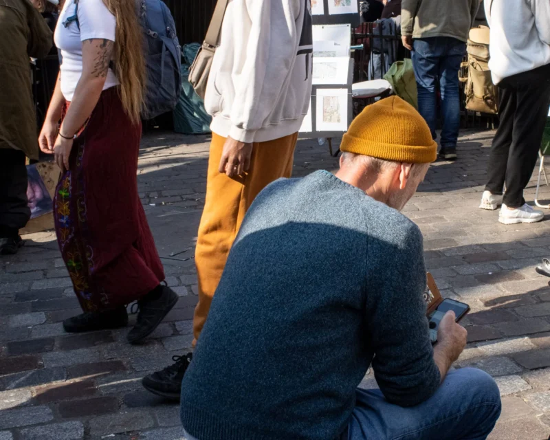 Loubna · 2048 x 1638 Cette photographie montre un homme assis comme sujet principal, concentré sur son téléphone. La composition est dynamique, avec des personnes en arrière-plan légèrement floues, créant une profondeur de champ.Le cadrage est serré sur l’homme, mettant en valeur ses détails vestimentaires (veste bleu-gris, chapeau jaune). La lumière semble naturelle, probablement en extérieur, avec des ombres douces. Les couleurs dominantes sont les tons froids (bleu, gris) et quelques touches chaudes (jaune du chapeau, rouge et marron des vêtements en arrière-plan).Le style est documentaire et spontané, évoquant une ambiance urbaine et détendue.