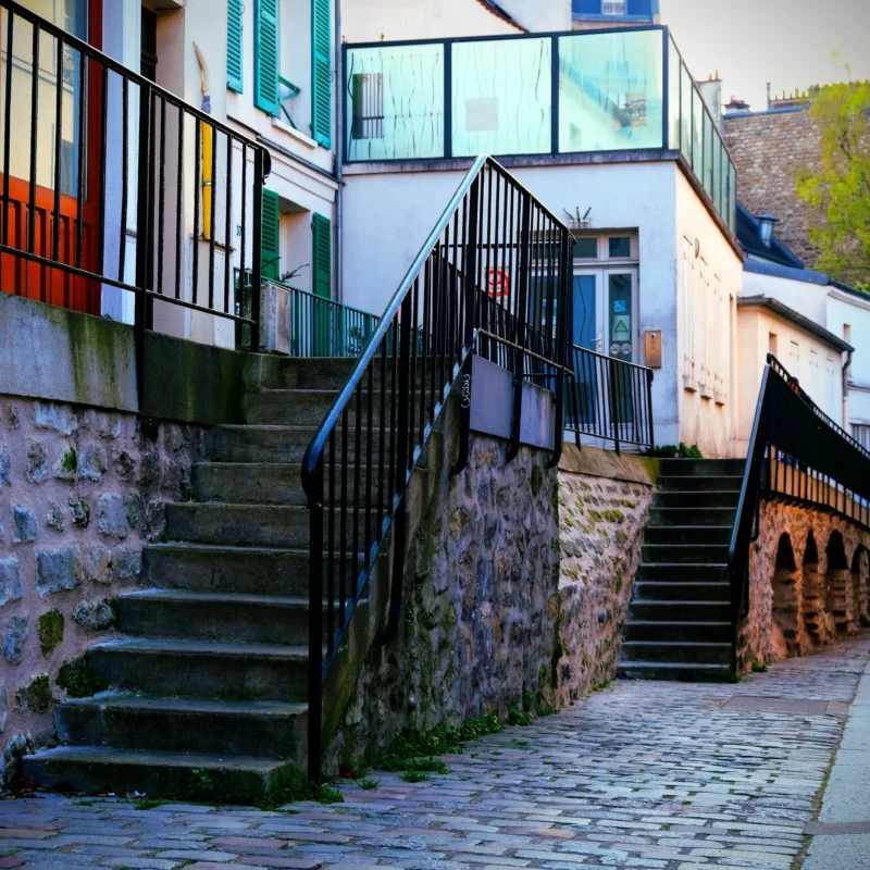 Denis · 21/03/2026 · 3265 x 3266 Cette photographie montre un escalier en pierre bordé de rampes métalliques menant à des maisons colorées. Le sujet principal est l’architecture résidentielle et les escaliers en pente.La composition est symétrique, avec deux jeux d'escaliers encadrant une allée pavée. Le cadrage est serré, mettant en valeur les détails architecturaux. La lumière est naturelle, douce et légèrement tamisée, créant des ombres douces.Les couleurs dominantes sont les tons pastel des façades (bleu, vert, rose) et les teintes grises des pierres et du métal. Le style est urbain et pittoresque, évoquant une ambiance chaleureuse et paisible.