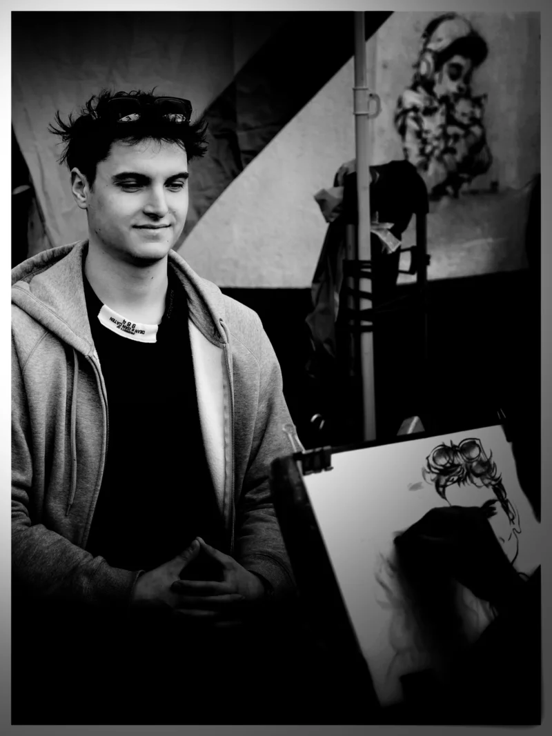 Denis · 22/03/2026 · 2177 x 2904 Cette photographie en noir et blanc met en scène un jeune homme comme sujet principal, souriant et tenant une planche à dessin. La composition montre un atelier artistique, avec des œuvres accrochées en arrière-plan. Le cadrage est serré sur le sujet, mais inclut des éléments contextuels.La lumière est douce, créant des contrastes modérés et mettant en valeur les traits du visage et les détails des dessins. Les couleurs dominantes sont les nuances de gris, typiques du noir et blanc.Le style est documentaire et artistique, évoquant une ambiance chaleureuse et créative, presque intimiste.
