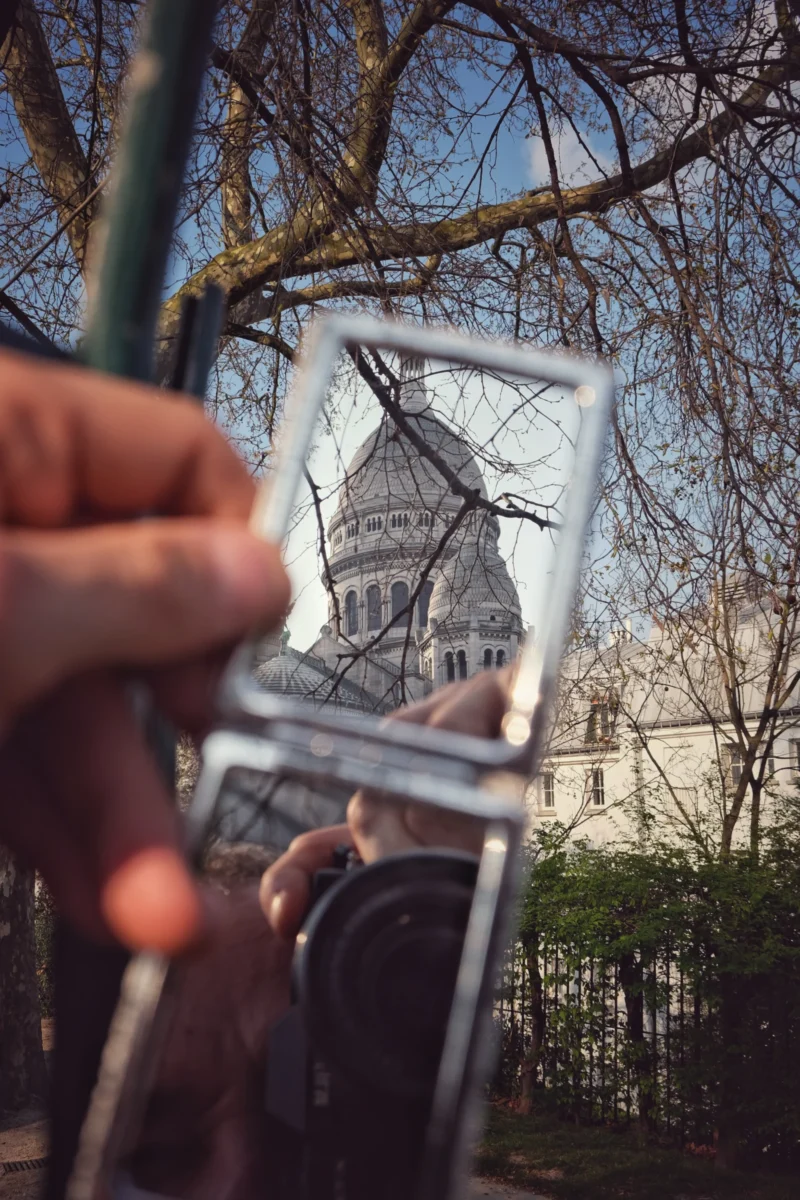Denis · 21/03/2026 · 3265 x 4898 Cette photographie montre une main tenant un miroir de poche reflétant une tour emblématique, probablement Big Ben. La composition joue sur le contraste entre le reflet et le cadre réel, avec des branches d’arbre en arrière-plan. Le cadrage est serré, mettant en valeur les détails du miroir et de la tour.La lumière est naturelle, douce et légèrement dorée, créant une ambiance claire et légèrement nostalgique. Les couleurs dominantes sont les tons gris-bleutés de la tour, les verts des feuilles et les teintes chaudes de la lumière. Le style est épuré et artistique, évoquant une touche de poésie et de mélancolie.