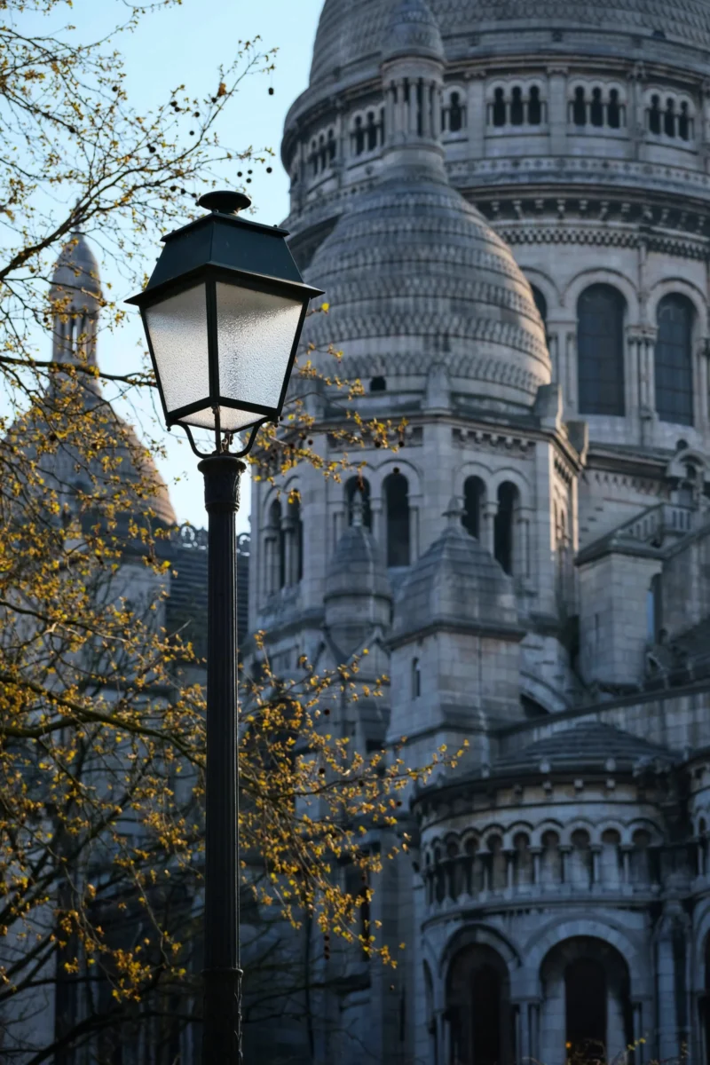 Denis · 21/03/2026 · 3265 x 4898 Cette photographie met en scène le **Sacré-Cœur de Montmartre** à Paris. Le **sujet principal** est la basilique, avec ses dômes et sa façade ornés de détails architecturaux.La **composition** est verticale, centrée sur une tour, avec un **cadrage serré** qui isole partiellement le bâtiment. Un **réverbère** et des **branches d’arbre** aux feuilles jaunes encadrent la scène.La **lumière** est douce, probablement crépusculaire, créant une ambiance paisible. Les **couleurs dominantes** sont les tons **beige et gris** de la pierre, contrastés par les **jaunes dorés** des feuilles.Le **style** est photographique classique, mettant en valeur l’architecture et l’atmosphère **romantique et sereine**.