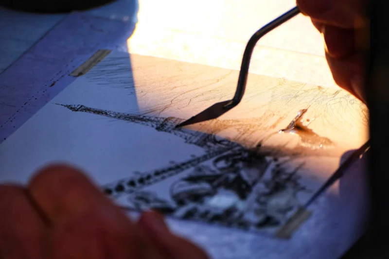 Denis · 21/03/2026 · 4898 x 3265 Cette photographie montre une personne en train de dessiner ou d’annoter un plan ou une carte à l’aide d’un stylo-plume. Le sujet principal est les mains et le travail minutieux sur le papier.La composition est serrée, centrée sur les détails du dessin. Le cadrage est rapproché, mettant en valeur les traits et les textures. La lumière douce, probablement naturelle, éclaire la scène en créant des contrastes subtils. Les couleurs dominantes sont des tons neutres (beiges, gris) avec des touches de bleu et de blanc.Le style est réaliste et précis, évoquant une ambiance studieuse et concentrée.