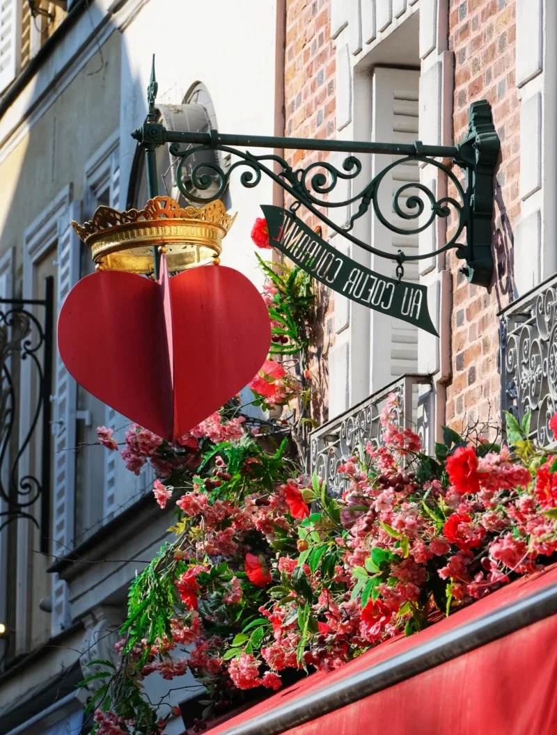 Denis · 22/03/2026 · 3265 x 4307 Cette photographie met en scène un balcon parisien orné de manière romantique. Le sujet principal est un cœur rouge suspendu à une balustrade en fer forgé, décorée de fleurs rouges et vertes. La composition est symétrique et harmonieuse, avec un cadrage serré sur le balcon.La lumière naturelle éclaire la scène, mettant en valeur les détails. Les couleurs dominantes sont le rouge vif des fleurs et du cœur, contrastant avec le noir de la balustrade et le vert des feuilles. Le style évoque un charme romantique et traditionnel, typique des décors parisiens. L’ambiance est chaleureuse et poétique.