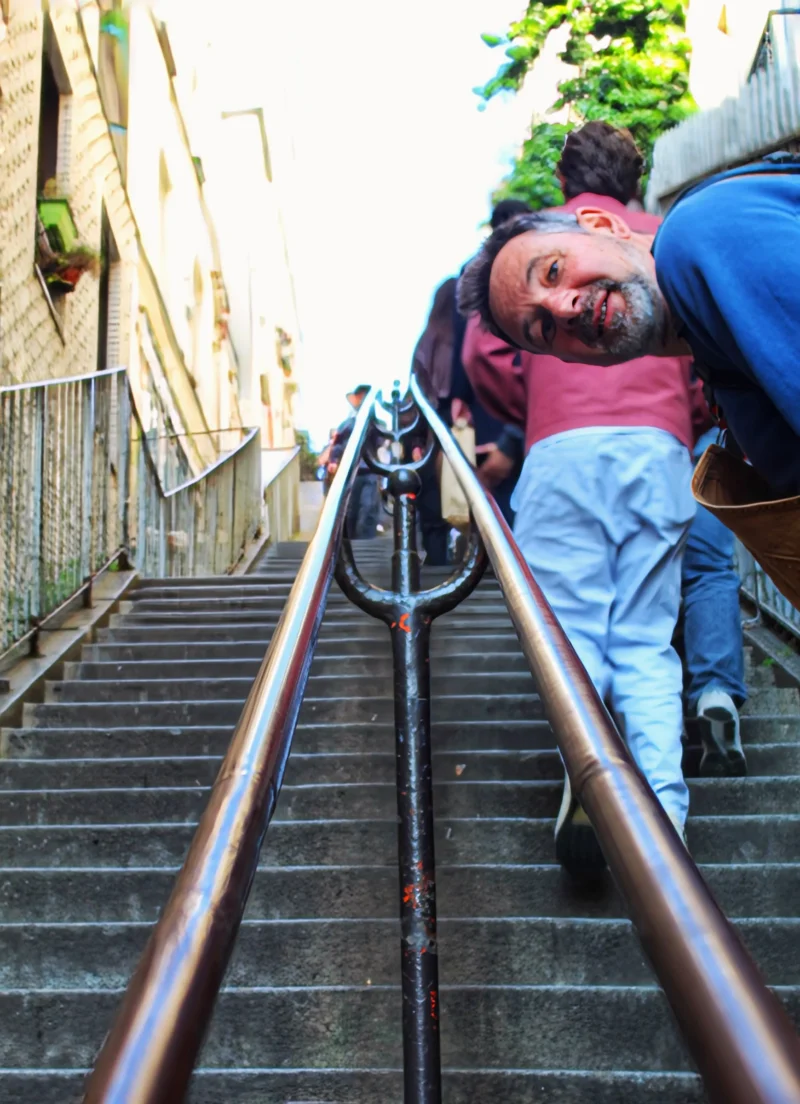 Vartika · 18/02/2026 · 3456 x 4771 Cette photographie montre un funiculaire en pente raide avec deux personnes assises sur les rails. Le sujet principal est le visage souriant et expressif d’un homme en premier plan, encadré par les rails métalliques.La composition est dynamique, avec un cadrage serré sur les rails et les passagers. La lumière est naturelle, légèrement contrastée, mettant en valeur les détails des visages et des rails. Les couleurs dominantes sont les tons gris des rails, les teintes vives des vêtements (rose, bleu) et les nuances naturelles des murs.Le style est documentaire et spontané, créant une ambiance joyeuse et insolite.