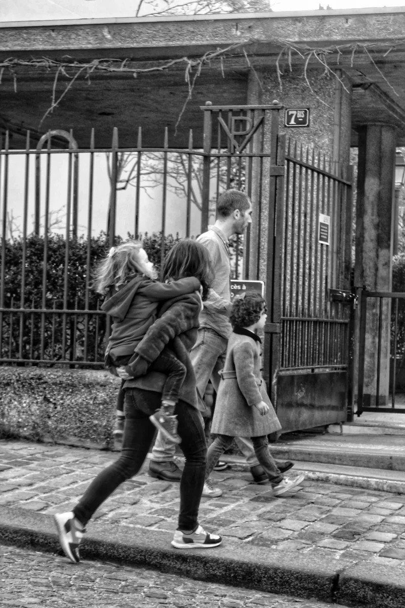Vartika · 18/02/2026 · 3456 x 5184 Cette photographie en noir et blanc capture un groupe de trois personnes, dont deux enfants et un adulte, traversant une rue pavée. Le sujet principal semble être les enfants, l’un courant devant, l’autre en mouvement avec l’adulte.La composition est dynamique, avec un cadrage serré qui met en valeur les sujets en mouvement. La lumière naturelle, douce mais contrastée, accentue les ombres et les contours. L’ambiance est spontanée et joyeuse, évoquant une scène de vie quotidienne des années 1960-70. Les couleurs, absentes en raison du noir et blanc, laissent place à des nuances de gris, renforçant le côté rétro et authentique.