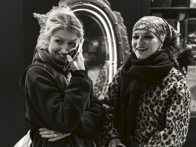 Portraits de rue Cette photographie en noir et blanc met en scène deux femmes comme sujets principaux. Elles sont positionnées côte à côte, l’une légèrement en arrière-plan près d’un miroir, créant une composition symétrique et réfléchie.Le cadrage est serré, mettant l’accent sur leurs visages et leurs tenues élégantes. La lumière est douce, avec des contrastes modérés, accentuant les détails des vêtements et des expressions.Les couleurs dominantes, bien que monochromes, suggèrent des tons sombres et des textures variées. Le style évoque une ambiance rétro ou vintage, renforcée par les accessoires et coiffures. L’ambiance est à la fois sophistiquée et intime.