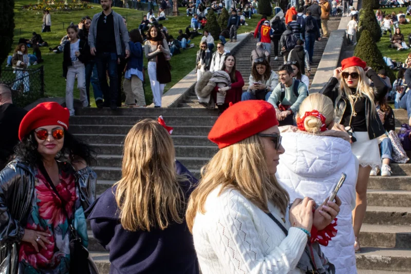 Loubna · 2048 x 1365 Cette photographie capture un groupe de personnes en extérieur, probablement lors d’un événement festif. Le sujet principal semble être les trois femmes en premier plan, vêtues de tenues colorées et coiffées de chapeaux rouges, évoquant une ambiance folklorique ou carnavalesque.La composition montre un escalier en pierre avec une foule en arrière-plan, créant une profondeur. Le cadrage est serré sur les femmes, mais inclut aussi des éléments contextuels. La lumière est naturelle et claire, suggérant une journée ensoleillée. Les couleurs dominantes sont vives : rouge, blanc, et des teintes pastel, renforçant l’ambiance joyeuse et festive. Le style est documentaire et spontané, reflétant une scène authentique et animée.