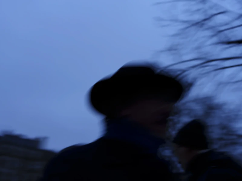 Une nuit dans la Marais Cette photographie présente un sujet principal flou, probablement une personne portant un chapeau, suggérant un mouvement. La composition est simple, avec une silhouette peu distincte. Le cadrage est serré, centré sur le visage et le haut du corps.La lumière est tamisée, créant une ambiance crépusculaire ou nocturne. Les couleurs dominantes sont les bleus froids et les teintes sombres, renforçant une atmosphère mystérieuse et mélancolique. Le style est épuré et artistique, privilégiant l’émotion à la précision. L’ambiance évoque le calme et une certaine introspection.