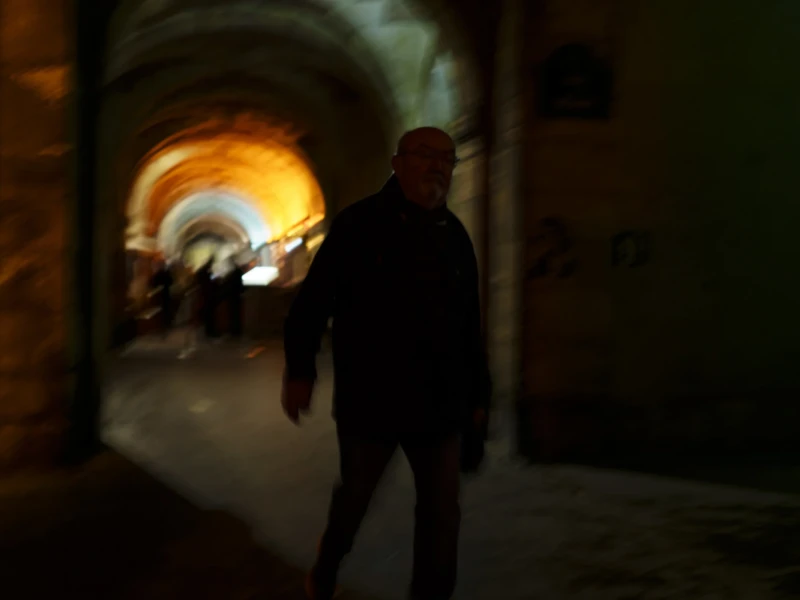Place des Vosges Cette photographie met en scène un homme de dos, silhouette principale dans un couloir sombre. La composition joue sur le contraste entre la lumière vive au fond, éclairant une arche, et l’obscurité environnante. Le cadrage serré et légèrement flou isole le sujet, créant une ambiance mystérieuse et intime.Les couleurs dominantes sont les tons chauds (jaunes, oranges) au centre, opposés aux teintes sombres (noirs, gris) en périphérie. Le style est réaliste, avec une touche de flou artistique. L’ambiance est à la fois dramatique et contemplative, évoquant solitude et profondeur.