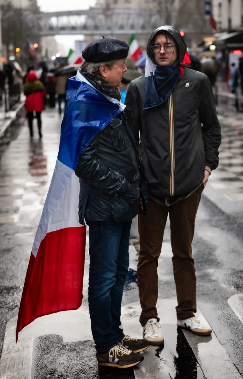 Pascal SC · 01/02/2026 · 1230 x 1920 Cette photographie capture deux personnes marchant sous une pluie légère, l’une portant un drapeau français. Le sujet principal est cette personne avec le drapeau, soulignant un possible engagement politique ou patriotique. La composition est dynamique, avec un cadrage serré sur les deux individus, mettant en valeur leurs expressions et leurs vêtements.La lumière est naturelle mais tamisée par la pluie, créant une ambiance sobre. Les couleurs dominantes sont les tons froids (gris, bleu) et les couleurs vives du drapeau (bleu, blanc, rouge). Le style est documentaire et réaliste, évoquant une scène authentique et spontanée, renforçant une ambiance à la fois sobre et émouvante.