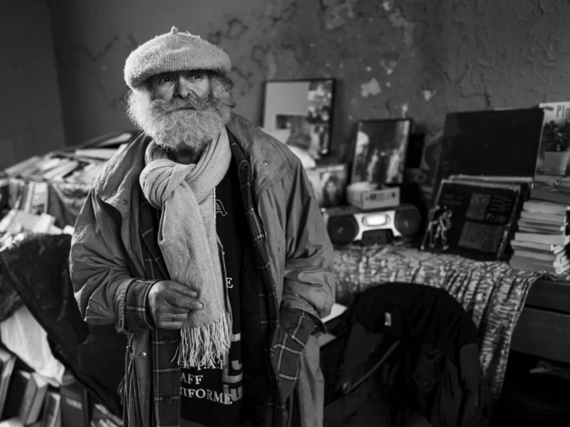 Portrait de rue Cette photographie en noir et blanc met en scène un vieil homme comme sujet principal, vêtu d’un manteau usé et d’un béret, tenant une bouteille. La composition montre un intérieur modeste, rempli d’objets divers (livres, vêtements, couvertures).Le cadrage est serré, centré sur lui, avec un arrière-plan légèrement flou. La lumière est douce, créant des contrastes modérés et mettant en valeur les textures. Les couleurs dominantes sont les nuances de gris, renforçant une ambiance mélancolique et réaliste.Le style évoque un documentaire social, avec une touche d’humanité et de simplicité. L’ambiance est sobre et introspective.