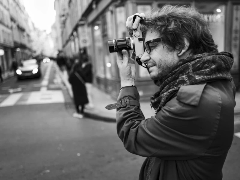 Thomas de Photos in Paris Cette photographie met en scène un homme comme sujet principal, tenant une caméra à l'œil, prêt à prendre une photo. La composition est simple et centrée sur lui, avec une rue urbaine en arrière-plan légèrement flou.Le cadrage est serré, mettant l'accent sur son visage et ses gestes. La lumière est douce et naturelle, typique d’un éclairage urbain crépusculaire ou nocturne. Les couleurs dominantes sont en noir et blanc, renforçant l’aspect dramatique et intemporel.Le style est documentaire et réaliste, évoquant une ambiance contemplative et artistique dans un cadre urbain animé.