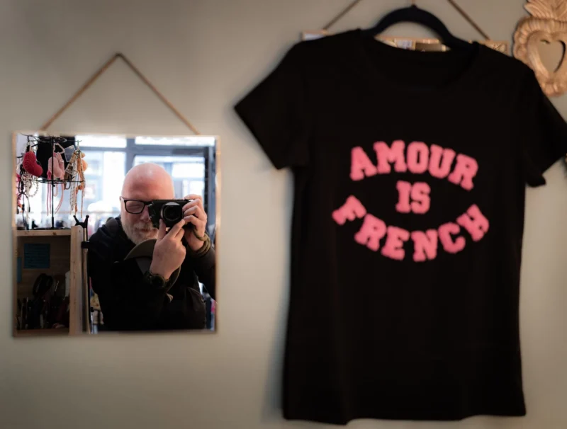 Pascal SC · 21/03/2026 · 1920 x 1455 Cette photographie montre un t-shirt noir accroché avec l'inscription "Amour is French" en lettres roses. À gauche, un miroir reflète une personne prenant une photo avec un appareil argenté.La composition est simple et symétrique, avec un cadrage serré sur les éléments principaux. La lumière semble naturelle et douce, éclairant uniformément la scène. Les couleurs dominantes sont le noir du t-shirt, le rose des lettres, et les tons neutres du fond.Le style est minimaliste et décontracté, évoquant une ambiance intime et quotidienne.