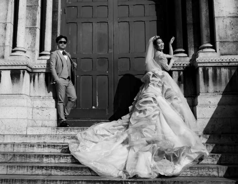 Pascal SC · 21/03/2026 · 1920 x 1482 Cette photographie en noir et blanc met en scène un couple élégant devant un escalier monumental. La femme, vêtue d'une longue robe de mariée traditionnelle, est le sujet principal, en mouvement descendant les marches. L’homme, en costume sobre, l’observe avec un chapeau.La composition joue sur les lignes architecturales et la symétrie. Le cadrage serré sur eux crée une ambiance intime malgré le décor imposant. La lumière douce, probablement naturelle, accentue les contrastes et les textures. Le style classique et romantique évoque une scène à la fois solennelle et poétique, renforcée par le monochrome.