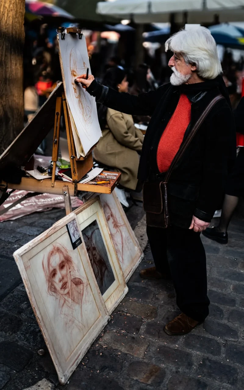 Pascal SC · 21/03/2026 · 1199 x 1920 Cette photographie montre un vieil homme, sujet principal, tenant une planche à dessin et observant ses œuvres. La composition met en avant ses peintures posées au sol et sur des chevalets. Le cadrage est serré, centré sur lui et ses tableaux.La lumière semble naturelle, légèrement tamisée, créant des ombres douces. Les couleurs dominantes sont sobres : tons beiges, gris et rouges, avec des touches de noir et blanc dans les dessins.L’ambiance est intimiste et artistique, évoquant un marché ou une rue animée, avec une touche de mélancolie et de créativité.