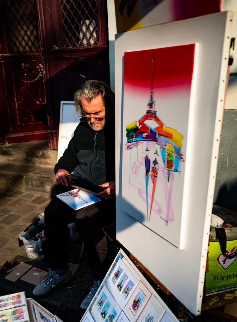 Pascal SC · 21/03/2026 · 1412 x 1920 Cette photographie montre un homme assis devant une grande toile illustrée, représentant une silhouette stylisée devant la Tour Eiffel. Le sujet principal est cet artiste, probablement en train de travailler ou de présenter son œuvre.La composition est dynamique, avec un cadrage serré sur l’artiste et son tableau. La lumière est naturelle et semble provenir de la gauche, créant des contrastes doux. Les couleurs dominantes sont vives : rouge, bleu, jaune et noir, typiques d’un style pop art ou street art.L’ambiance est artistique et créative, évoquant un marché ou une exposition de rue.