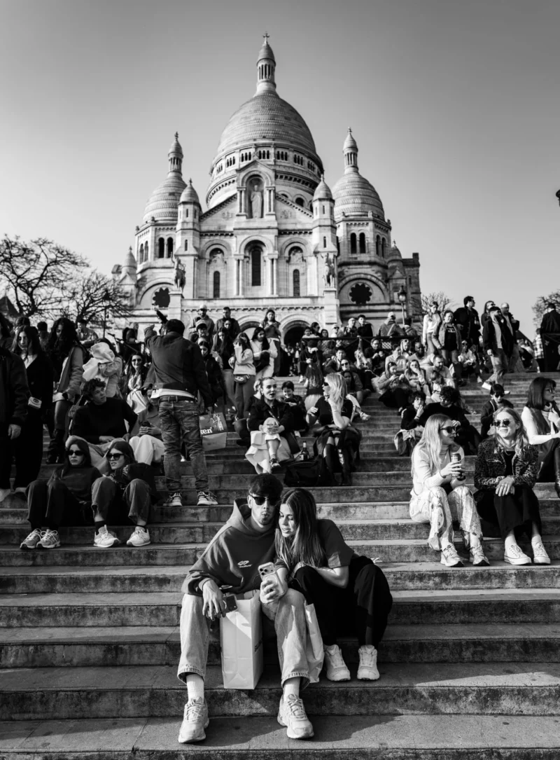 Pascal SC · 21/03/2026 · 1416 x 1920 Cette photographie en noir et blanc montre le Sacré-Cœur de Paris comme sujet principal. La composition met en avant les marches remplies de monde, avec un groupe assis au premier plan. Le cadrage est serré sur les personnes, intégrant partiellement l’architecture emblématique en arrière-plan.La lumière est naturelle, douce et diffuse, créant des contrastes subtils. L’absence de couleurs accentue les textures et les jeux d’ombres. Le style est documentaire et réaliste, capturant une scène quotidienne. L’ambiance est à la fois animée et paisible, reflétant une vie sociale et touristique intense.
