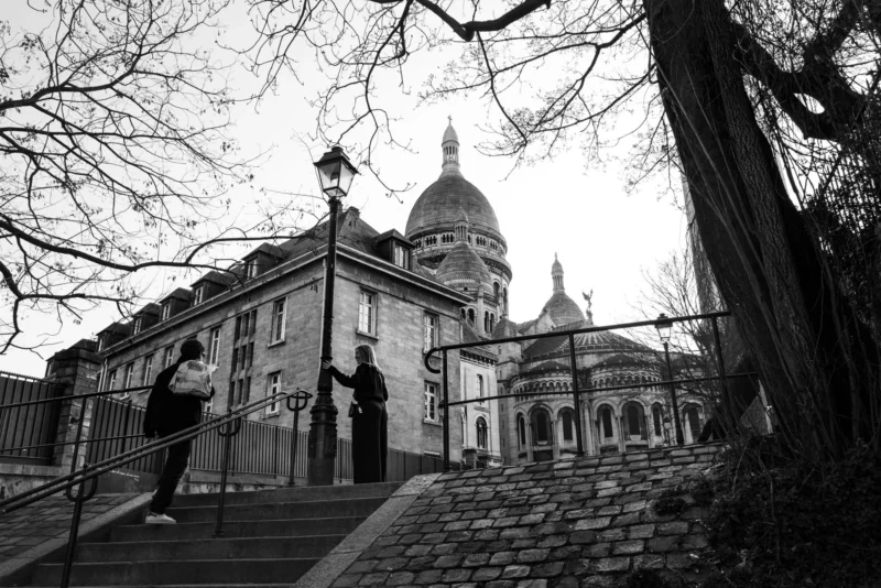 Pascal SC · 21/03/2026 · 1920 x 1281 Cette photographie en noir et blanc met en scène un couple descendant un escalier devant un bâtiment historique, probablement une cathédrale ou un édifice religieux, reconnaissable à sa flèche et ses détails architecturaux.La composition est symétrique, avec les marches menant vers le centre de l’image. Le cadrage inclut des arbres dénudés et une lanterne, ajoutant une touche de profondeur. La lumière est douce, créant des contrastes subtils entre ombres et clartés. Les couleurs dominantes sont les tons de gris, renforçant une ambiance sobre et intemporelle. Le style est classique et élégant, évoquant une scène à la fois romantique et solennelle.