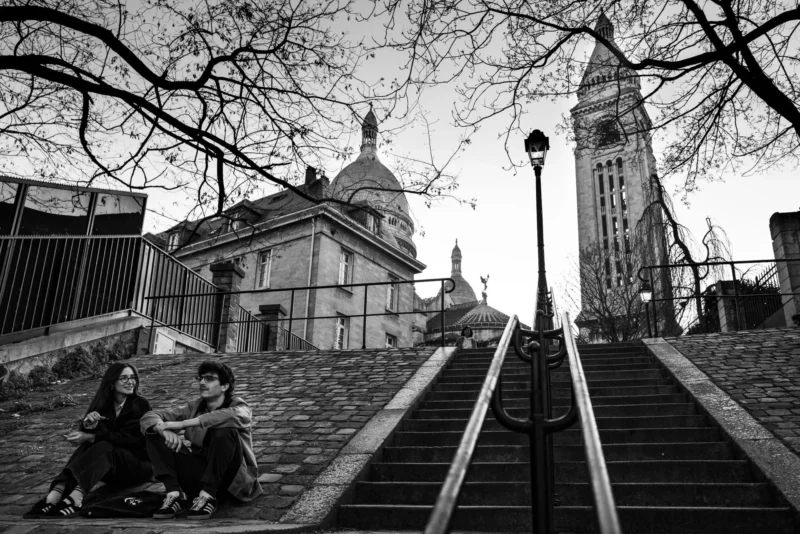 Pascal SC · 21/03/2026 · 1920 x 1281 Cette photographie en noir et blanc met en scène deux personnes assises sur des marches, formant le sujet principal. La composition joue sur la perspective avec une montée d'escaliers menant vers un monument et une église en arrière-plan.Le cadrage est serré sur les deux individus, tout en intégrant un large champ de profondeur. La lumière est douce, avec des contrastes marqués créant des ombres nettes. Les couleurs dominantes, bien que monochromes, exploitent les nuances de gris pour accentuer les textures et les formes.Le style est documentaire et intemporel, évoquant une ambiance mélancolique et contemplative.