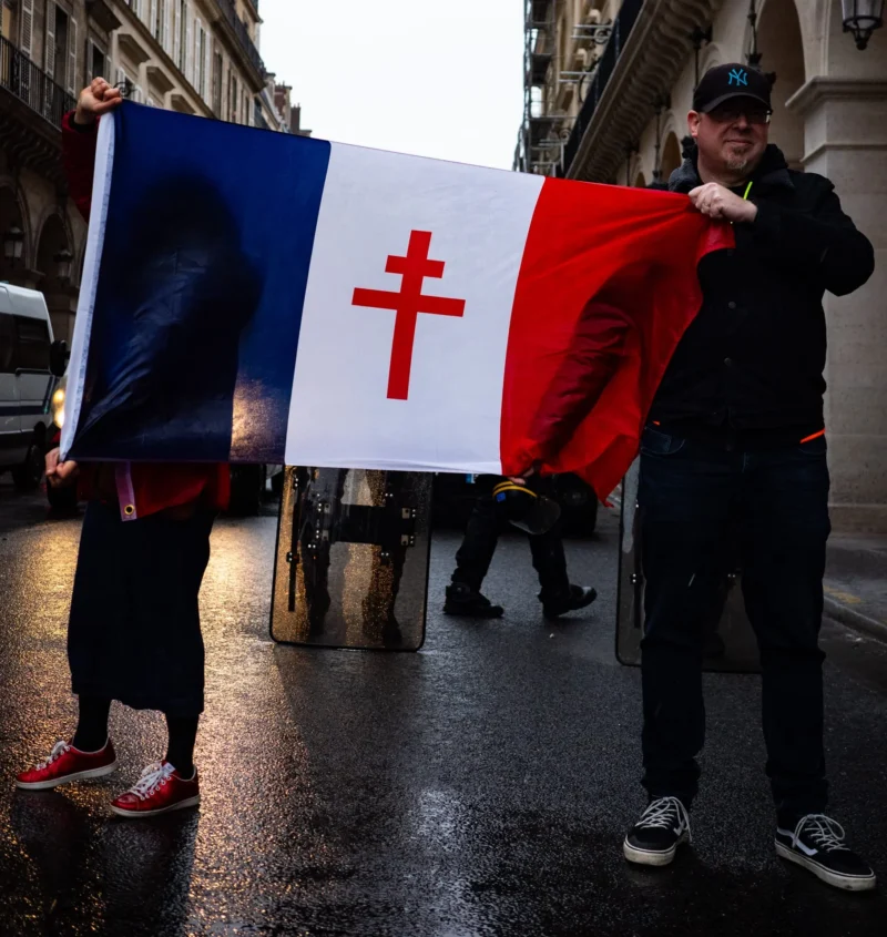 Pascal SC · 22/02/2026 · 1817 x 1920 Cette photographie montre deux personnes tenant un drapeau aux couleurs bleu, blanc et rouge avec une croix rouge stylisée, évoquant la France. Le sujet principal est ce drapeau, symbole de protestation ou de soutien.La composition est dynamique, avec un cadrage serré sur les individus et leur drapeau. L’ambiance est urbaine, sous une lumière naturelle grise et diffuse, typique d’un jour pluvieux. Les couleurs dominantes sont les tons froids (bleu, gris) et les contrastes vifs du drapeau. Le style est documentaire, capturant un moment authentique et engagé. L’ambiance est à la fois sérieuse et mobilisatrice.