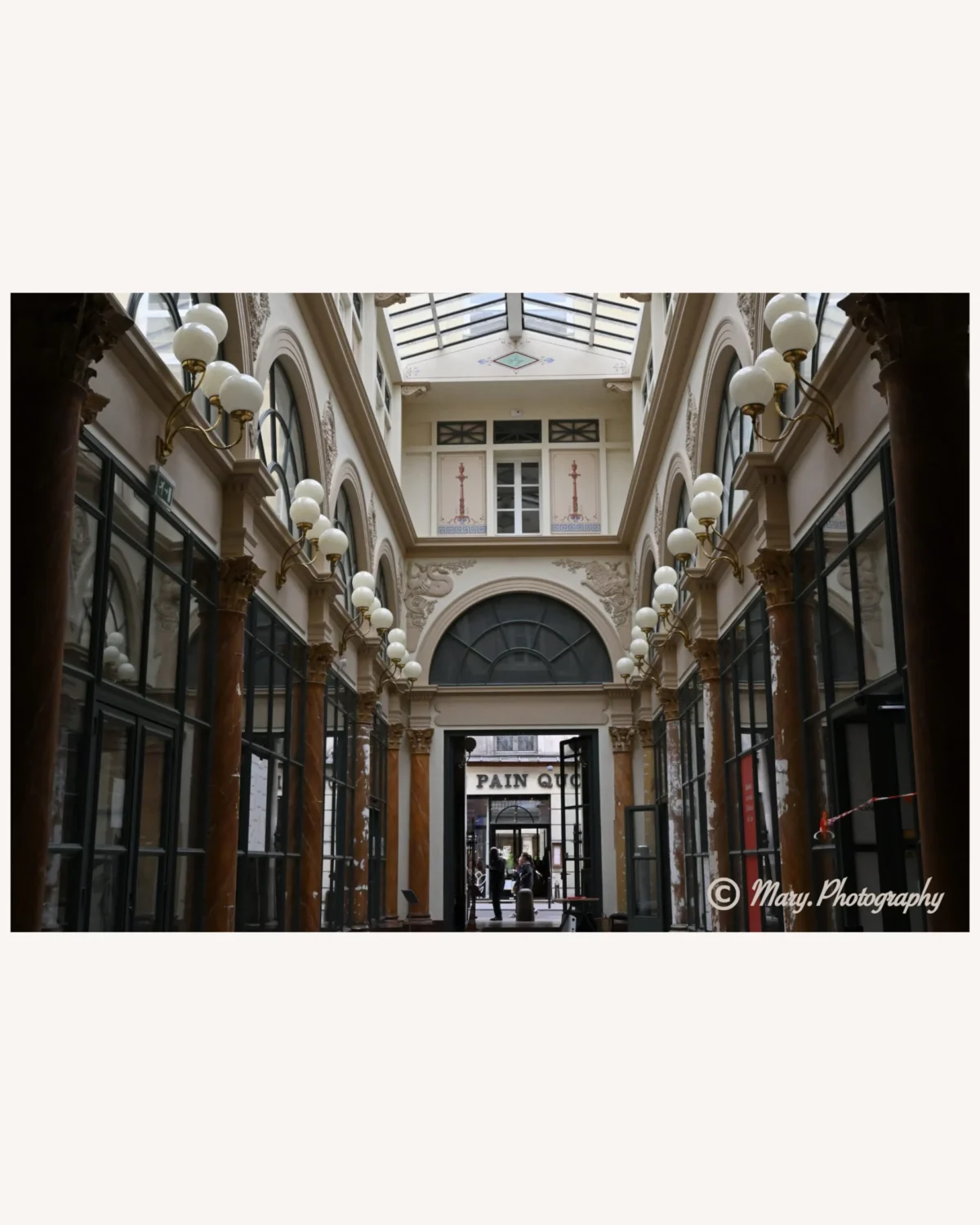 Mary · 2048 x 2560 Cette photographie représente une galerie marchande couverte avec une architecture classique. Le sujet principal est l'alignement des colonnes et des vitrines, encadrant une perspective profonde.La composition utilise un cadrage serré et symétrique, créant une sensation de profondeur et de continuité. La lumière est douce et naturelle, filtrée par les vitraux du plafond, éclairant uniformément l'espace.Les couleurs dominantes sont des tons neutres : beige, gris et marron, avec des touches de noir et de blanc. Le style est élégant et intemporel, évoquant une ambiance raffinée et paisible, presque nostalgique.