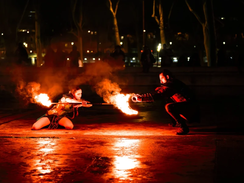 Burners du Palais de Tokyo Cette photographie capture une scène nocturne de danse ou performance avec des éléments de feu. Le sujet principal est un danseur en mouvement, entouré de flammes tenues par d'autres participants.La composition est dynamique, avec un cadrage serré sur les artistes. Les lumières sont chaudes et contrastées, mettant en valeur les flammes et créant une ambiance dramatique. Les couleurs dominantes sont les tons rouges et oranges des feux, contrastant avec les teintes sombres de la nuit. Le style semble artistique et culturel, évoquant une ambiance mystique et intense.