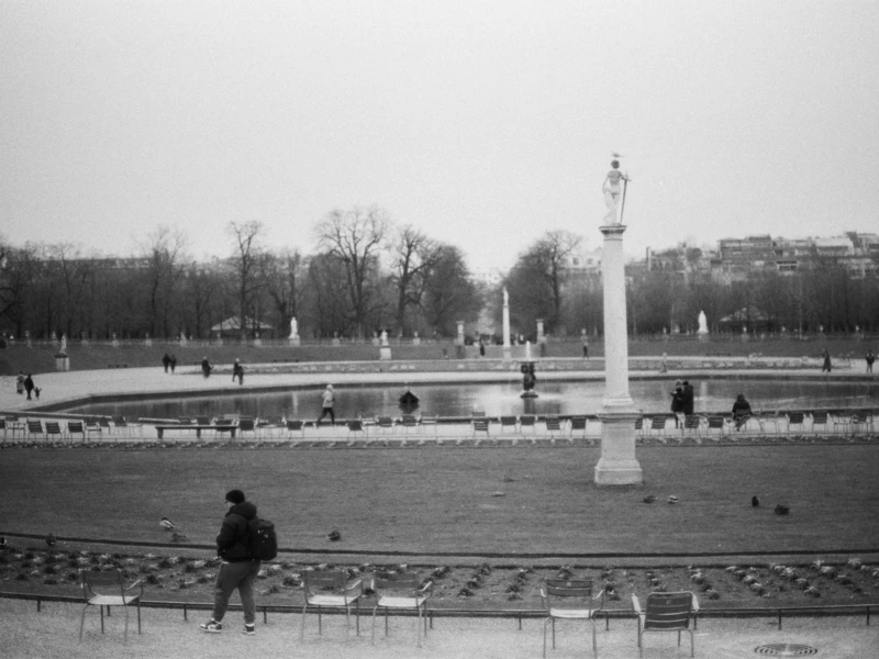 Cette photographie en noir et blanc montre un parc urbain, probablement les Jardins du Luxembourg à Paris. Le sujet principal est une colonne centrale entourée de grilles et de statues.La composition est symétrique, avec des allées et des arbres alignés. Le cadrage est large, incluant une personne assise à gauche. La lumière est douce, typique d’un jour couvert, adoucissant les contrastes. Les couleurs dominantes, en noir et blanc, accentuent les textures et les ombres.Le style est documentaire et classique, évoquant une ambiance calme et intemporelle, presque mélancolique.