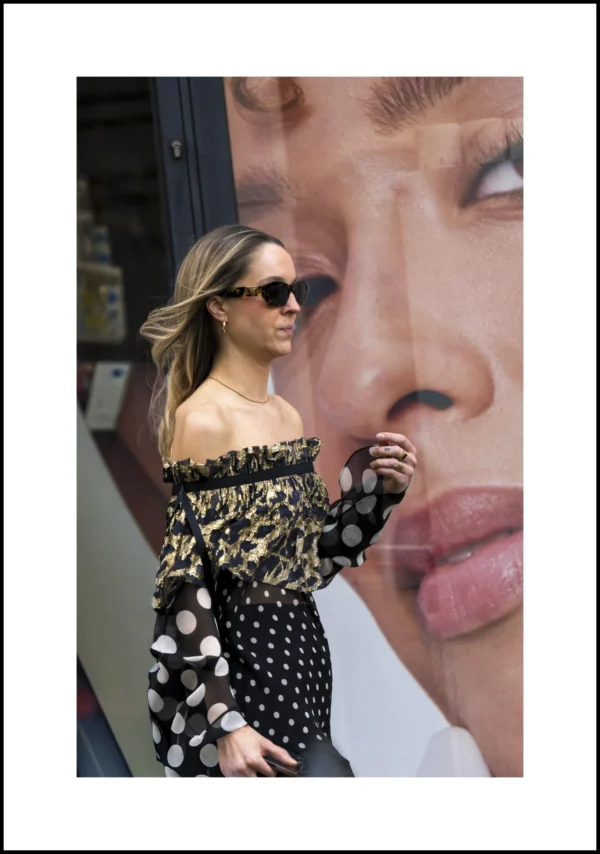 Thierry · 06/03/2026 · 3290 x 4680 Cette photographie met en scène une femme comme sujet principal, vêtue d’une robe à off-shoulder à pois noirs et blancs, portant des lunettes de soleil. Elle se tient devant une affiche géante représentant un visage féminin aux traits exagérés.La composition joue sur le contraste entre la personne réelle et son reflet déformé. Le cadrage est serré sur la femme, avec un arrière-plan partiel. La lumière semble naturelle, bien que contrastée. Les couleurs dominantes sont les noirs, blancs et gris, créant une ambiance à la fois élégante et surréaliste. Le style évoque un mélange de street style et d’art visuel.