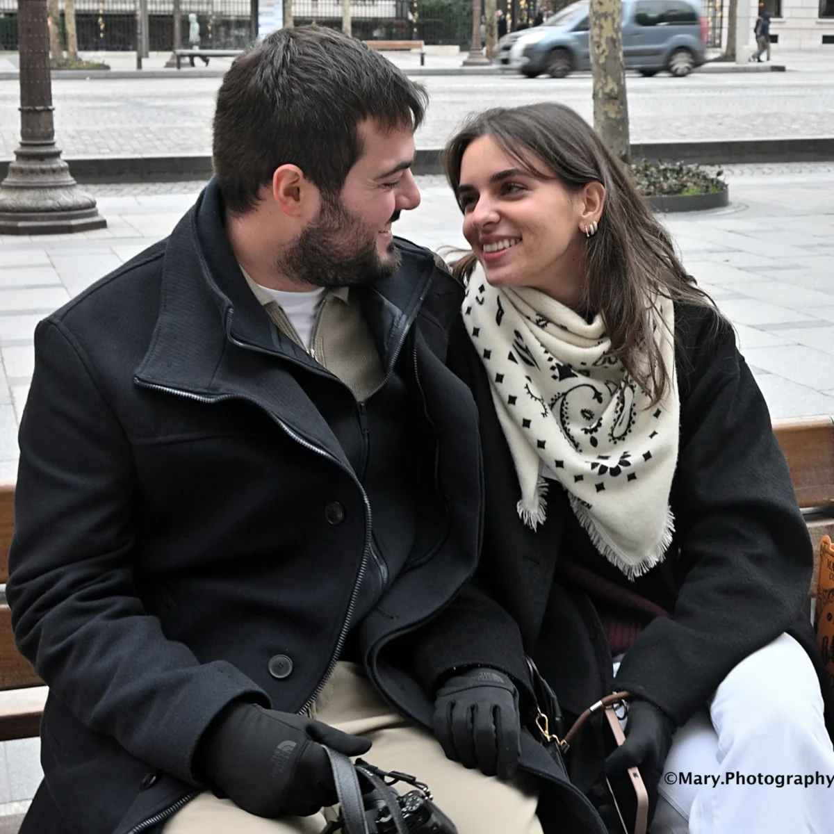 Mary · 2784 x 2784 Cette photographie met en scène un couple assis en extérieur. Le sujet principal est les deux personnes souriantes, partageant un moment complice. La composition est serrée, centrée sur eux, avec un cadrage rapproché. La lumière semble naturelle, douce et légèrement diffuse, typique d’un jour nuageux.Les couleurs dominantes sont sobres : tons gris (vêtements), noir (manteau de l’homme), et blanc (écharpe). L’ambiance est chaleureuse et intime, évoquant une connexion entre les deux individus. Le style est simple et spontané, proche du reportage.