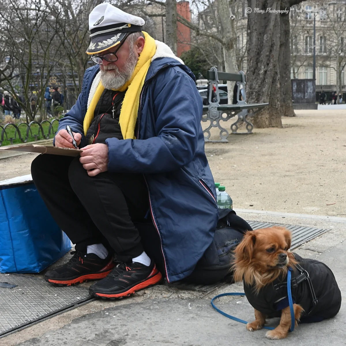 Mary · 2784 x 2784 Cette photographie met en scène un homme âgé comme sujet principal, assis sur un banc avec son petit chien. La composition est simple et naturelle, centrée sur eux deux.Le cadrage est serré, mettant en valeur leurs expressions et détails. La lumière semble naturelle, probablement en journée, avec des ombres douces. Les couleurs dominantes sont les tons neutres (gris du béton, bleu du manteau) et quelques touches chaudes (jaune du foulard, marron du chien).Le style est documentaire et réaliste, capturant un moment de vie quotidienne. L’ambiance est calme et touchante, évoquant la sérénité et la complicité entre l’homme et son animal.