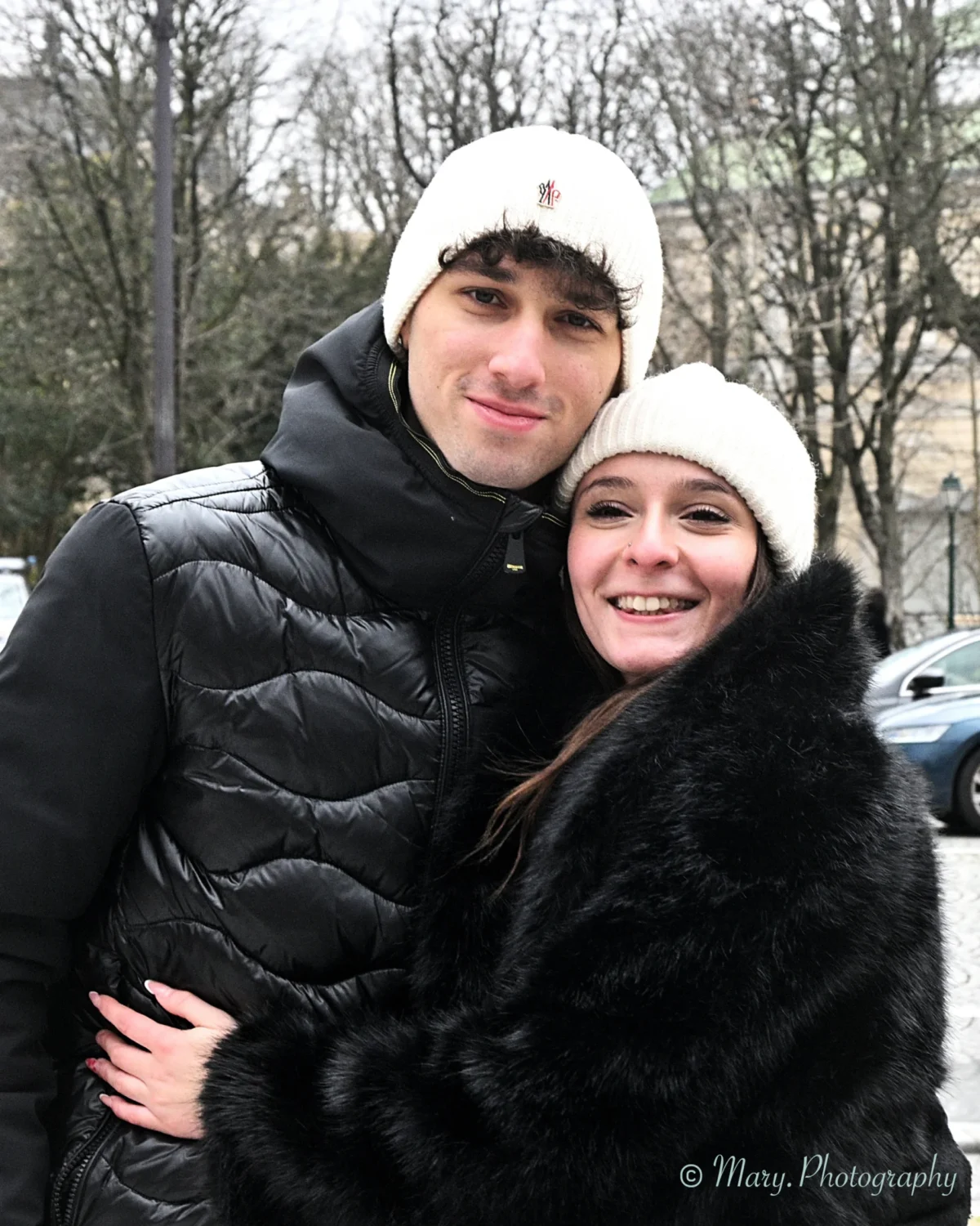 Mary · 2227 x 2784 Cette photographie met en scène un couple en hiver. Les sujets principaux sont un homme et une femme souriants, serrés l’un contre l’autre.Le cadrage est serré, centré sur eux. La composition est équilibrée, avec un arrière-plan flou de voitures et d’arbres.La lumière semble naturelle, douce, probablement en journée. Les couleurs dominantes sont les tons froids : noir (vêtements), gris (arrière-plan), et blanc (bonnets), avec une touche de fourrure noire.Le style est simple et spontané, évoquant une ambiance chaleureuse et intime malgré le froid.