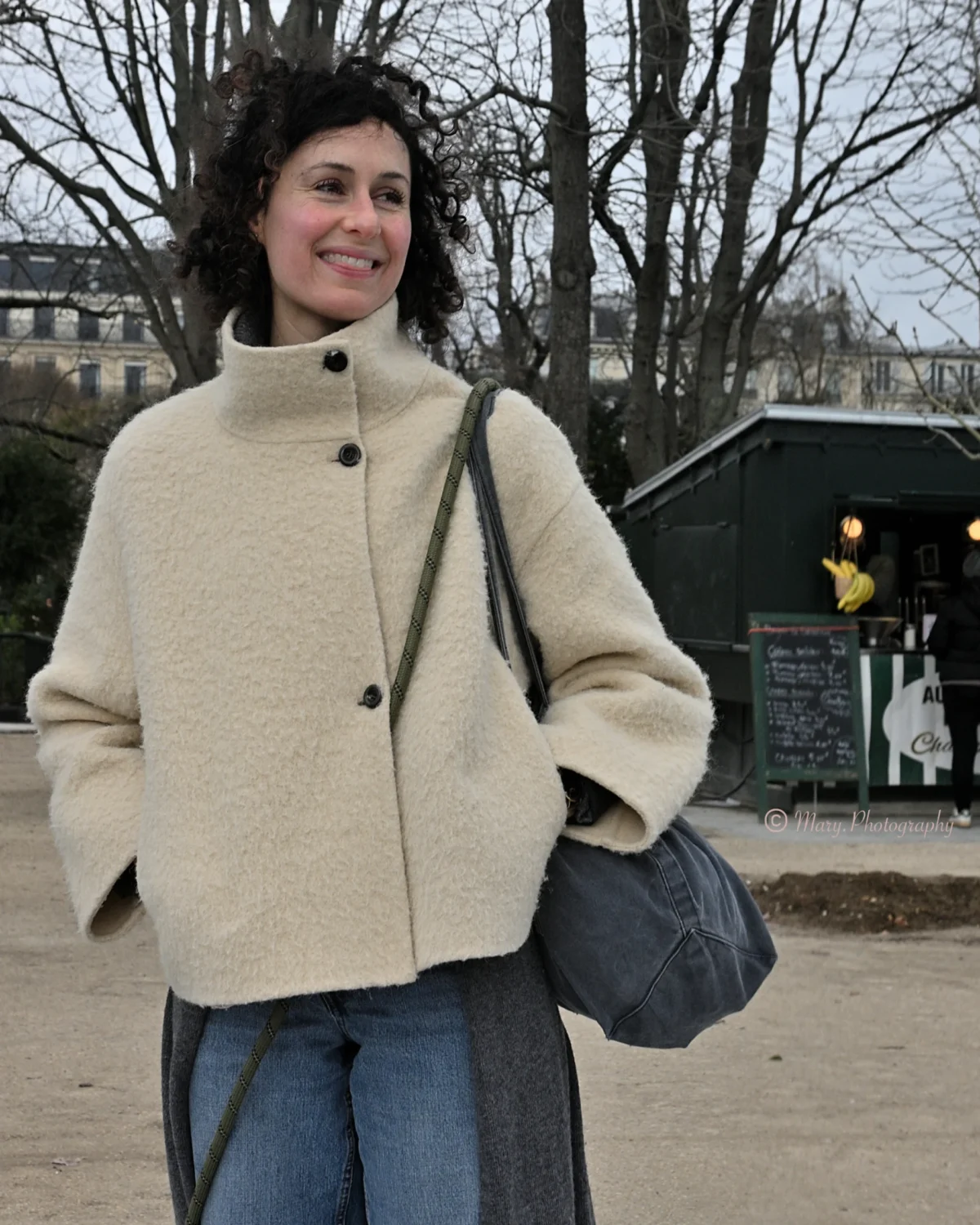Mary · 2227 x 2784 Cette photographie met en scène une femme souriante comme sujet principal, vêtue d’un manteau beige et d’un jean, avec un sac à main.La composition est simple et naturelle, centrée sur elle. Le cadrage est serré, incluant un arrière-plan flou avec des arbres et une structure urbaine.La lumière est douce et naturelle, probablement en journée. Les couleurs dominantes sont les tons beiges, bleus et verts discrets.Le style est décontracté et élégant, évoquant une ambiance urbaine et détendue, typique d’une promenade en ville.