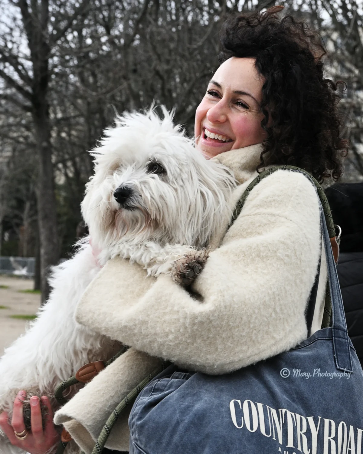 Mary · 2227 x 2784 Cette photographie met en scène une femme souriante tenant un petit chien blanc à poils longs. Le sujet principal est le duo, créant une ambiance chaleureuse et affectueuse.La composition est simple et centrée, avec la femme assise sur un banc. Le cadrage est serré, mettant en valeur les deux protagonistes. La lumière naturelle semble douce, probablement en journée, éclairant les visages et le pelage du chien.Les couleurs dominantes sont les tons neutres (beige, gris) et blancs, adoucis par le vert des arbres en arrière-plan. Le style est naturel et spontané, évoquant une scène de vie quotidienne paisible.