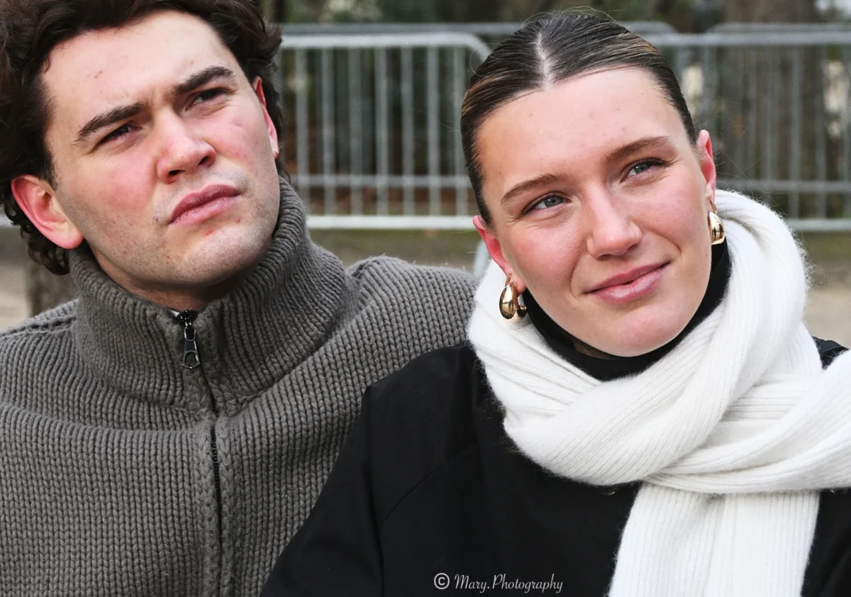 Mary · 2784 x 1952 Cette photographie met en scène deux jeunes posant côte à côte. Le sujet principal est leur expression et leur proximité. La composition est symétrique, avec un cadrage serré sur leurs visages et épaules.La lumière semble naturelle, douce et légèrement tamisée, créant des ombres subtiles. Les couleurs dominantes sont les tons neutres : gris du pull de l’homme, noir et blanc du vêtement de la femme, adoucis par le beige de son écharpe.Le style est sobre et réaliste, évoquant une ambiance intime et naturelle, presque quotidienne.