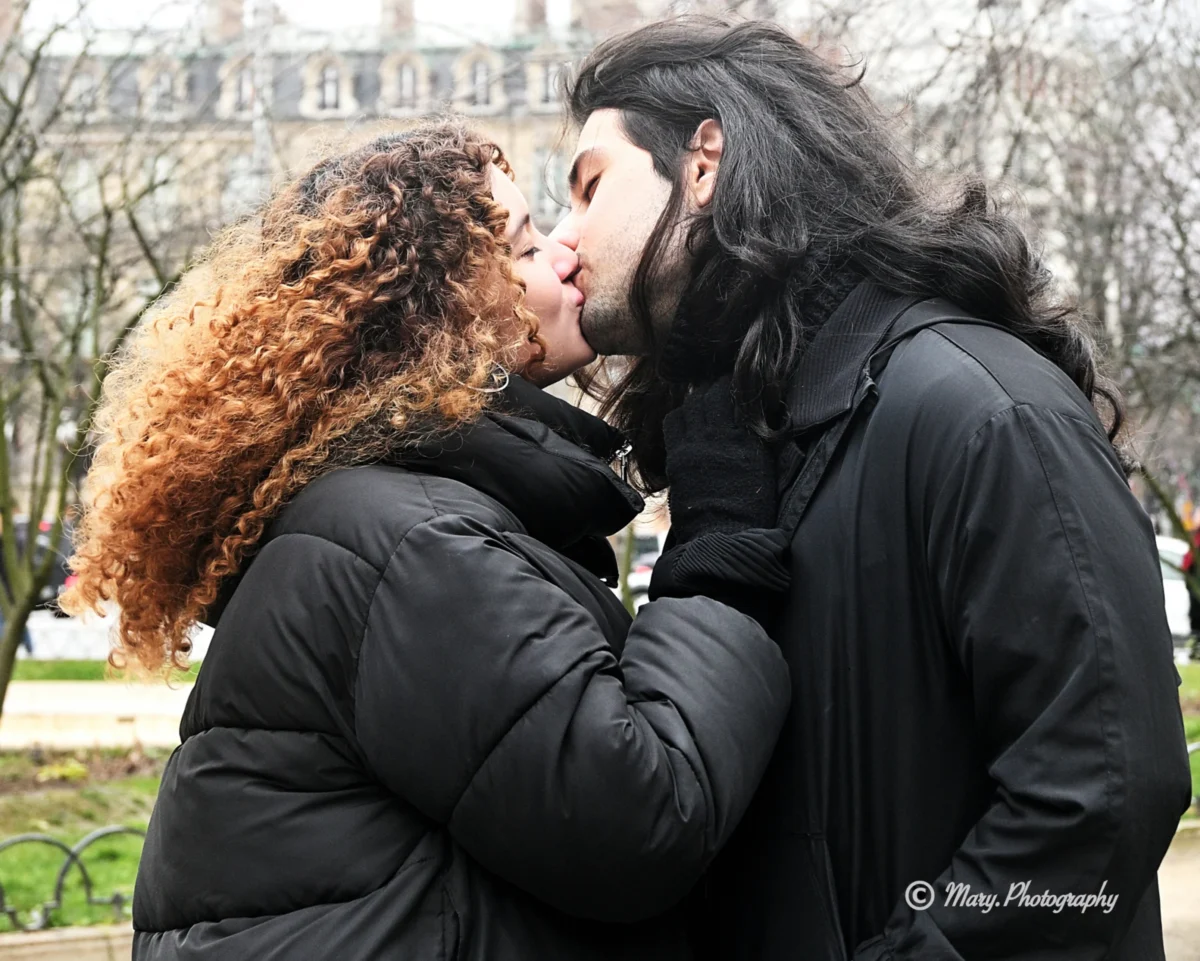 Mary · 2784 x 2228 Cette photographie met en scène un couple en train de s'embrasser tendrement. Le sujet principal est leur complicité et leur intimité.La composition est centrée sur eux, avec un cadrage serré qui isole les visages et les épaules. La lumière est douce et naturelle, probablement en journée, créant des ombres légères. Les couleurs dominantes sont les tons chauds des cheveux roux et les teintes neutres des vêtements, avec des touches de vert en arrière-plan.Le style est simple et romantique, évoquant une ambiance intime et affectueuse dans un cadre naturel et urbain.