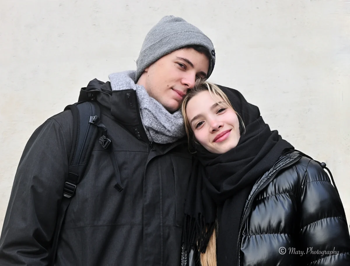 Mary · 2784 x 2117 Cette photographie met en scène un couple en position affectueuse. Le sujet principal est les deux jeunes, souriants et proches, créant une ambiance intimiste et chaleureuse.La composition est simple et centrée, avec un cadrage serré qui isole les visages et les épaules. La lumière semble naturelle et douce, probablement en extérieur par temps couvert, adoucissant les traits et évitant les ombres marquées.Les couleurs dominantes sont les tons froids : noir des vêtements, gris du fond et blanc du manteau du jeune homme, avec une touche de rouge apportée par les lèvres. Le style est épuré et spontané, évoquant une ambiance romantique et moderne.