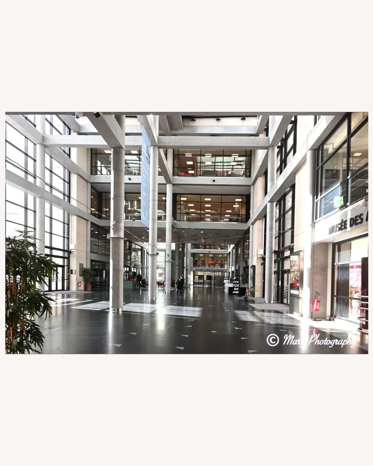 Mary · 2048 x 2560 Cette photographie montre un hall d’immeuble moderne. Le sujet principal est l’architecture intérieure avec ses grandes colonnes, ses escaliers et ses balcons vitrés. La composition est symétrique, centrée sur un espace ouvert et lumineux.Le cadrage est large, mettant en valeur les perspectives et les niveaux. La lumière naturelle, abondante, provient des grandes baies vitrées, créant une ambiance claire et aérée. Les couleurs dominantes sont neutres : gris, blanc et noir, avec des touches de bois clair. Le style est épuré et contemporain, évoquant une ambiance professionnelle et élégante.