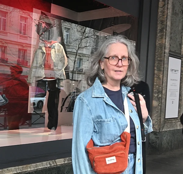 Mary P · 07/03/2026 · 2784 x 2633 Cette photographie montre une femme comme sujet principal, vêtue d'une veste bleue et portant un sac orange. Elle pose devant une vitrine affichant une œuvre artistique abstraite et des silhouettes.Le cadrage est serré sur elle, avec un arrière-plan partiellement visible. La lumière semble naturelle, bien répartie, sans ombres marquées. Les couleurs dominantes sont les tons bleus de la veste, l'orange du sac, et les teintes froides et contrastées de l'arrière-plan.Le style est simple et réaliste, évoquant une ambiance urbaine et détendue.