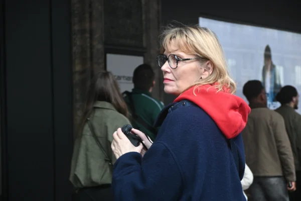 Mary P · 07/03/2026 · 5568 x 3712 Cette photographie met en scène une femme blonde, sujet principal, portant des lunettes et un vêtement bleu avec un gilet rouge. Elle semble observer son téléphone.La composition montre un arrière-plan flou avec plusieurs personnes, suggérant un lieu public. Le cadrage est serré sur elle, avec une légère profondeur de champ.La lumière est naturelle, probablement en extérieur, avec des ombres douces. Les couleurs dominantes sont les tons froids (bleu) et chauds (rouge). L’ambiance est sobre et quotidienne, évoquant une scène urbaine et informelle.